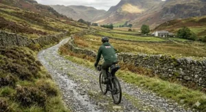 A cyclist navigating a wide, rocky bridleway on an electric bike packed with gear, heading towards a traditional stone cottage nestled in a sweeping British valley.