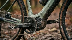 A macro photorealistic 4K photograph of a compact mid-drive electric motor unit (with a discrete logo) and mud-caked chainring, seamlessly integrated into the light green frame of the electric cyclocross bike seen in the featured image. The textures of dried mud, trail dust, and complex engineering are sharply in focus against a blurred forest background.