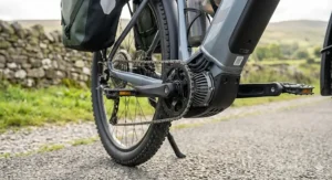 A detailed low-angle view of the mid-drive motor unit and crankset on a grey electric touring bicycle, highlighting the durable chain drive system against a dry stone wall background.