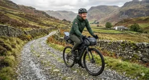 A side-angle shot of a smiling cyclist on a kitted-out electric gravel bike, showcasing frame bags and handlebar packs on a rugged, stone-walled trail in the UK countryside.
