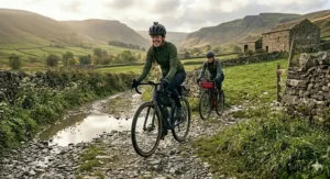 Two cyclists riding electric gravel bikes equipped with bikepacking bags down a wet, gravel track next to a classic dry stone wall and a stone outbuilding.