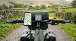Symmetric front-facing view of the electric touring bicycle cockpit, displaying ergonomic handlebar grips, a brass bell, a digital e-bike computer screen, and a smartphone mounted to the left showing a map routing through the British countryside.