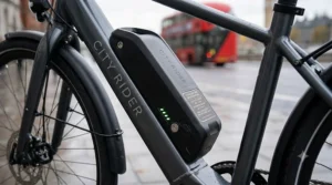 A detailed close-up illustration of the e-bike battery pack, featuring a specifications plaque detailing 504Wh capacity and 40-70 mile estimated range for the 250W motor system, placed against a blurred London street background with a red bus.