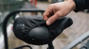 Hands stretching the elasticated hem of a weather-resistant e-bike saddle cover over a standard bicycle seat.